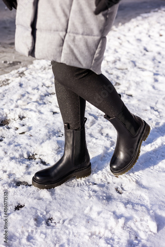 Woman in fashionable black boots on white snow, close up. Women's legs in stylish winter leather boots