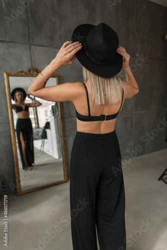 Glamorous fashionable young woman with sexy clothes in a black bra and pants with an elegant hat stands near the mirror with her back to the camera