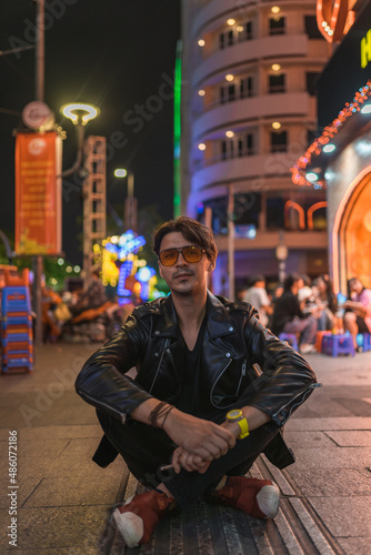 Portrait of young stylish brunet caucasian man in black leather jacket and yellow sunglasses Sitting on the street at night. High quality photo