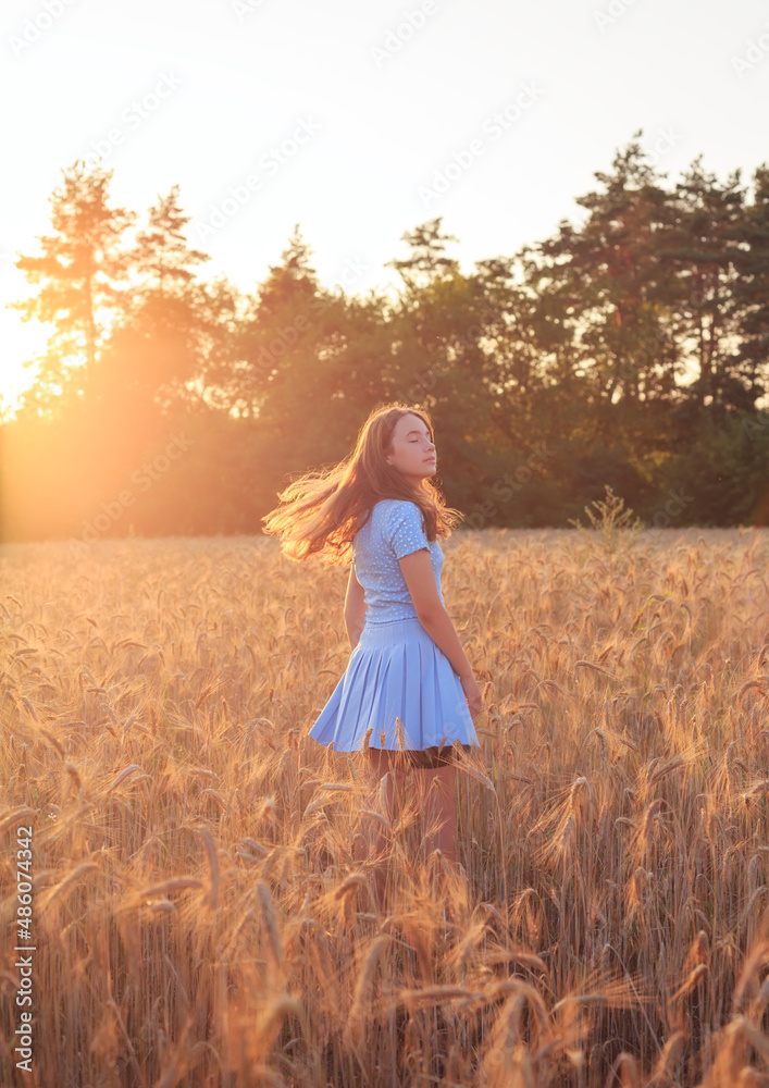 Obraz premium Happy teen girl is smiling at wheat field, touching ears of wheat with her hand. Beautiful teenager enjoying nature in warm sunshine in a wheat field on sunset background. Soft focused