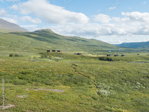 Northern landscape, tundra in Swedish Lapland with wooden cottages of STF Duottar tourist hut, blue artic river and lake, green hills and mountains at Padjelantaleden hiking trail. Summer day
