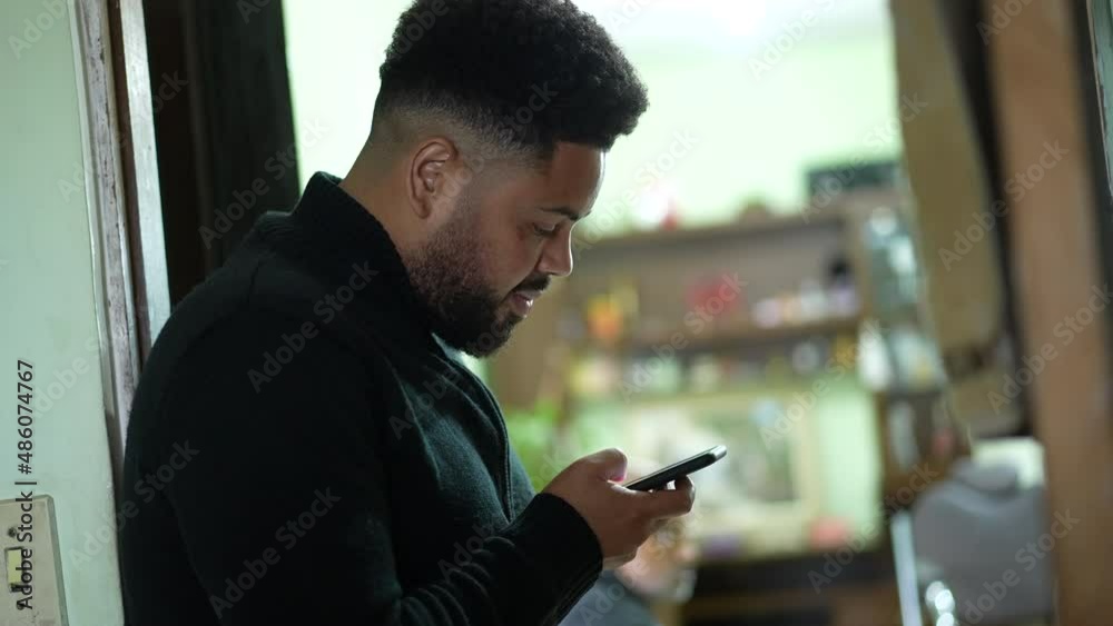 A hispanic man looking at cellphone device at home leaning on wall A hispanic man looking at cellphone device at home leaning on wall