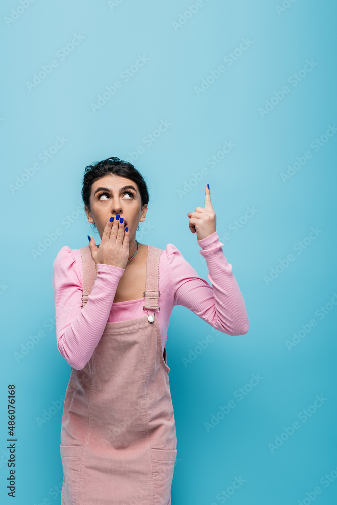 shocked woman looking up, pointing with finger and covering mouth with hand isolated on blue shocked woman looking up, pointing with finger and covering mouth with hand isolated on blue