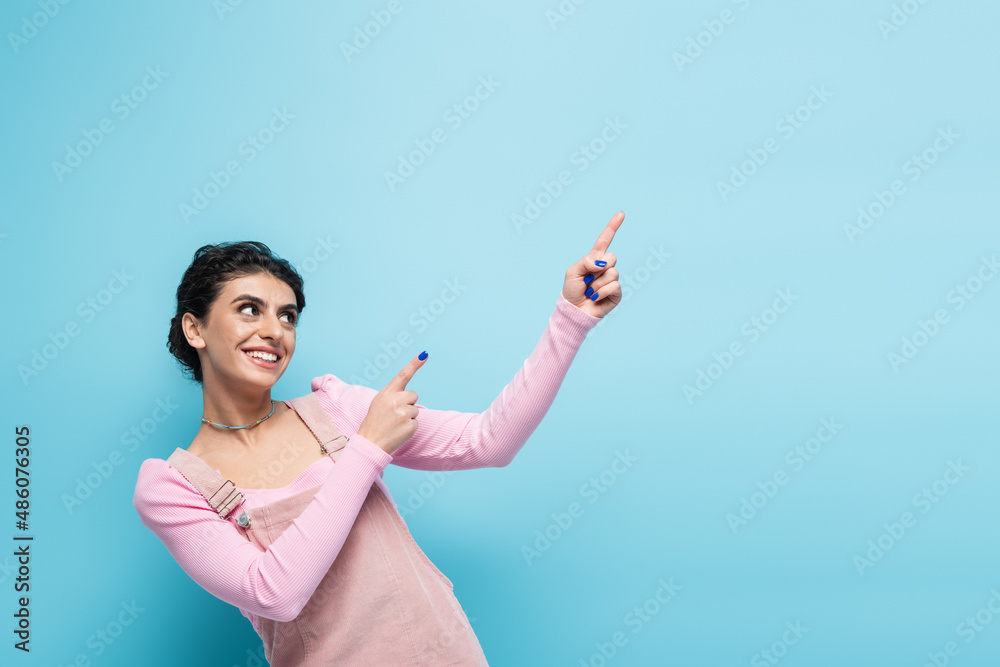 cheerful young woman in trendy pastel clothes looking away and pointing with fingers isolated on blue cheerful young woman in trendy pastel clothes looking away and pointing with fingers isolated on blue