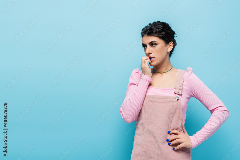 tense woman with hand near face looking away while standing with hand on hip isolated on blue tense woman with hand near face looking away while standing with hand on hip isolated on blue