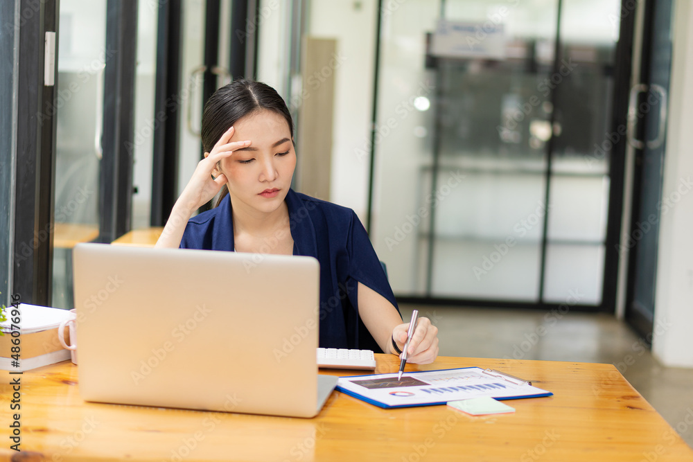 A young Asian woman accountant sits at her desk with her laptop and calculates financial graphs showing investment results. Plan a successful business growth process in the office. A young Asian woman accountant sits at her desk with her laptop and calculates financial graphs showing investment results. Plan a successful business growth process in the office.