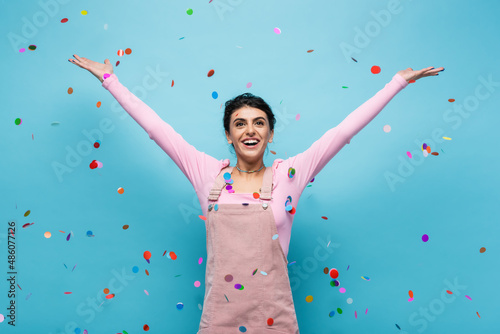 excited woman in pastel clothes smiling under falling confetti on blue background