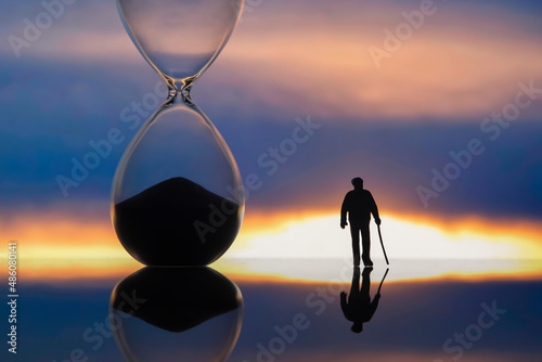 miniature people. silhouette of an elderly man walking towards the sunset next to an hourglass. end of life. © photosaint