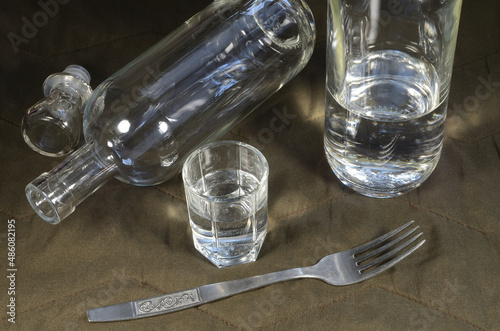 A bottle of vodka, a glass of vodka, a fork on a table with a brown background.
