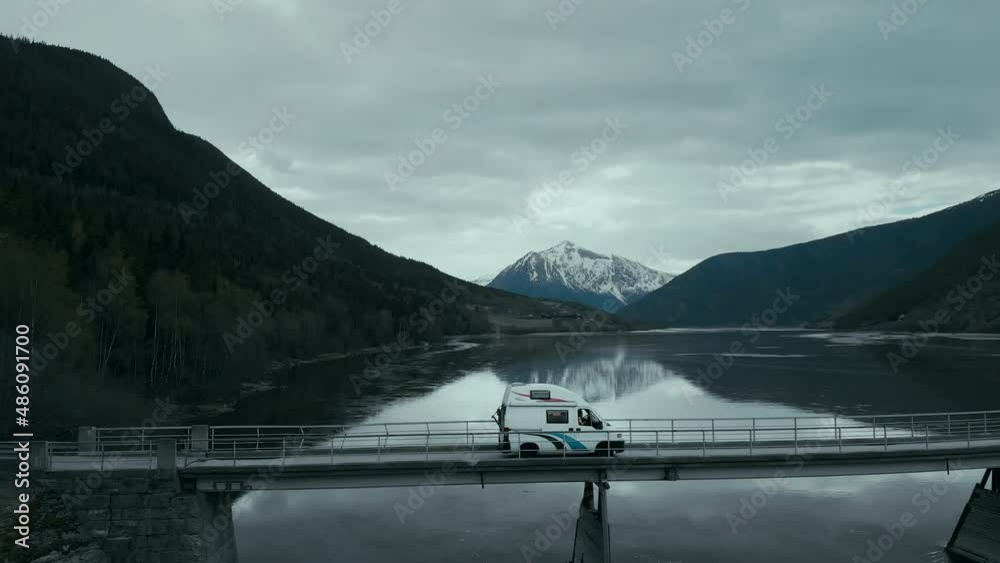 Incredible cinematic footage from drone point of view on cute vintage camper van driving on narrow bridge over fjord or mountain lake. Epic destination for nomad adventure camping lifestyle