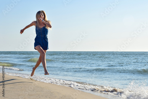 Young blond woman runs happy along the seashore beach in denim overalls