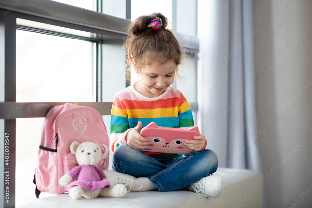 Positive little girl, holding tablet computer in her hands. Advertising new gadget, educational app. The girl sits near the window. She is wearing a colored T-shirt. Games.Children use technology. Positive little girl, holding tablet computer in her hands. Advertising new gadget, educational app. The girl sits near the window. She is wearing a colored T-shirt. Games.Children use technology.