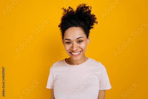 Portrait of happy joyful african american young woman with curly hair and freckles, wearing basic t-shirt, standing over isolated orange background, looking straight at camera, smiling friendly