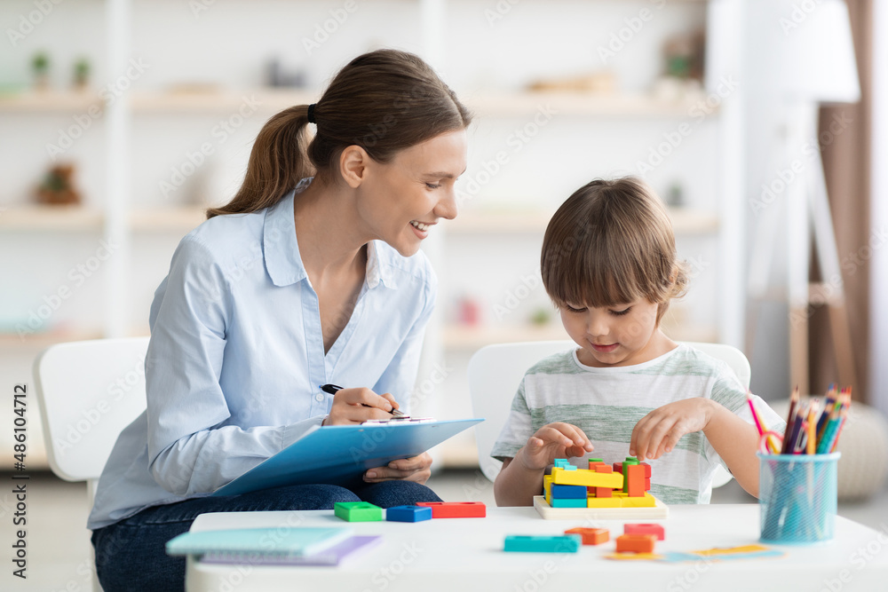 Early kids development. Cute little boy playing logical geometry game, woman specialist with notepad watching him Early kids development. Cute little boy playing logical geometry game, woman specialist with notepad watching him