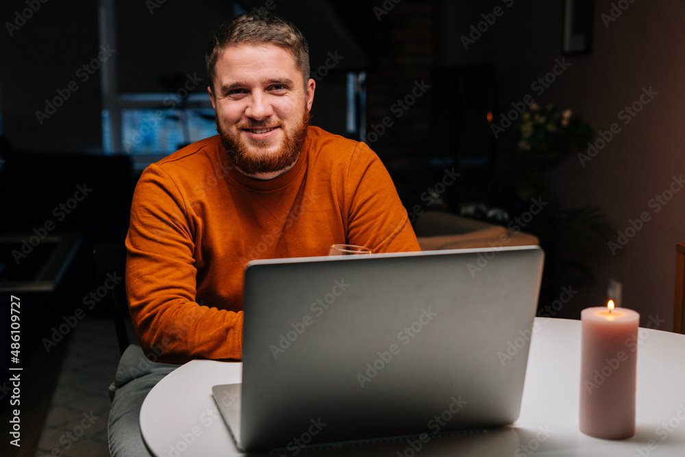 Smiling young man enjoying distant dating using laptop computer, holding glass with wine , talking celebration toasting via video call, sitting at table with candle in dark room, looking at camera. Smiling young man enjoying distant dating using laptop computer, holding glass with wine , talking celebration toasting via video call, sitting at table with candle in dark room, looking at camera.