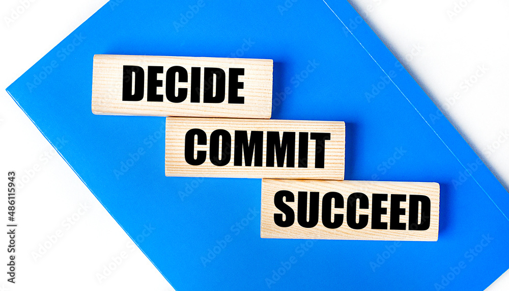 There is a blue notebook on a light gray background. Above are three wooden blocks with the words DECIDE COMMIT SUCCEED