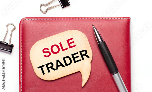 On a light background there are black paper clips, a pen, a burgundy notepad a wooden board with the text SOLE TRADER