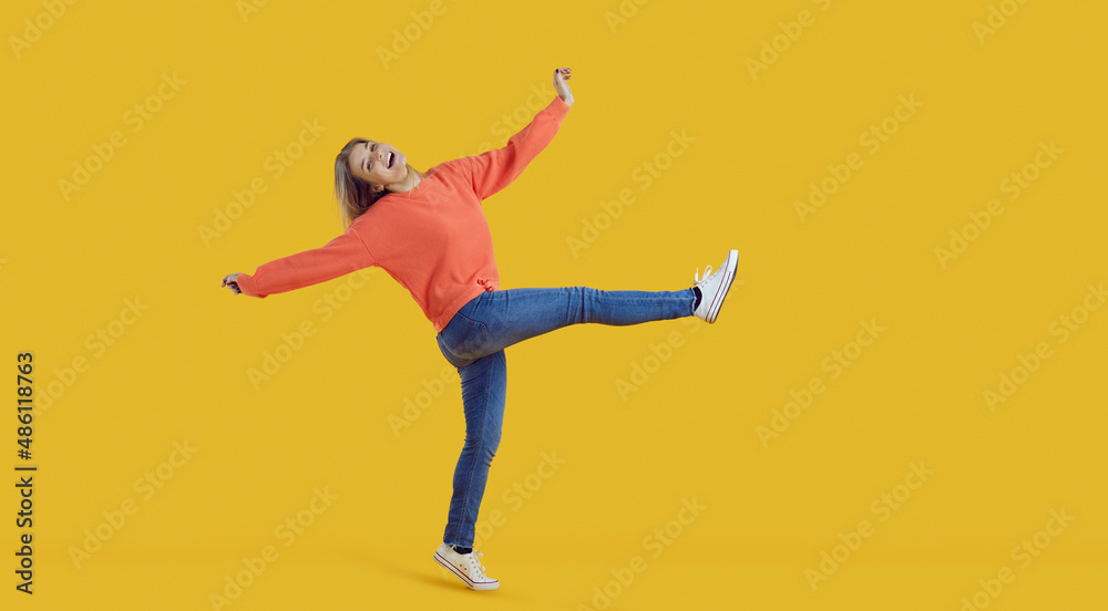 Funny carefree young woman in comfortable casual wear having fun and fooling around. Happy pretty teenage girl in orange sweatshirt and blue jeans dancing in studio with vibrant yellow background