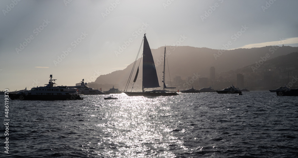 A lot of huge sail yachts and motor boats in port of Monaco at sunset, mountains are on background, megayachts are moored in sea, sun reflection on water, backlight