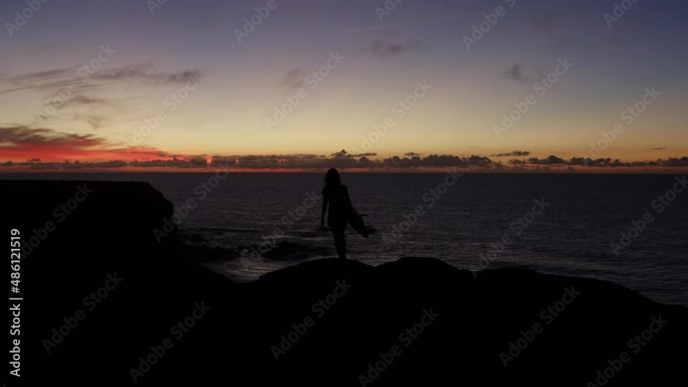 Aerial round shot of girl standing on a cliff with a surf in hand,looking at a beautiful sunset by the Atlantic ocean in Fuerteventura, Canary islands