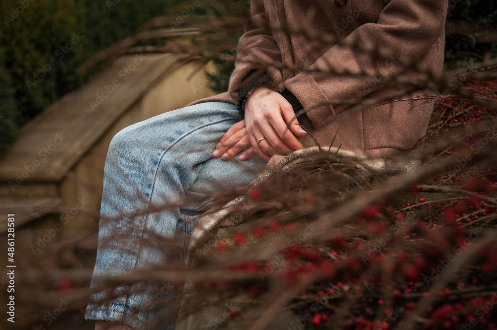 Fototapeta premium Woman in jeans and coat crossing hands sitting on plaid on steps railing in city park in early spring