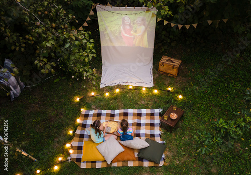 High angle view of children sitting on plaid and looking movie on backyard. Summer outdoor weekend activities with kids. Open air cinema.