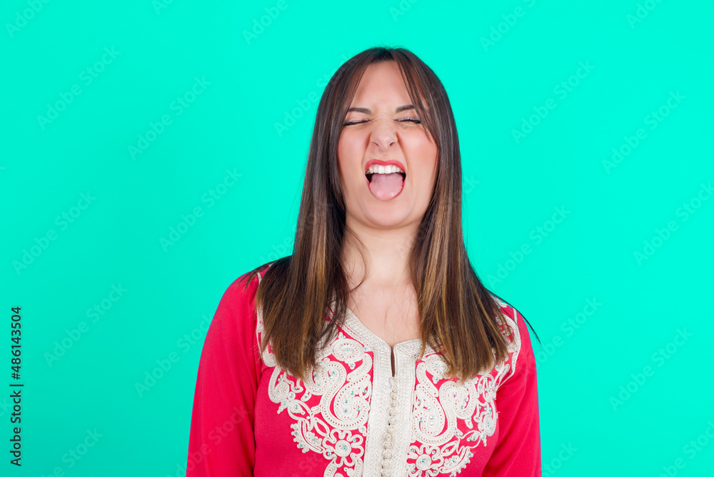 young beautiful moroccan woman wearing traditional caftan dress over green background sticking tongue out happy with funny expression. Emotion concept.