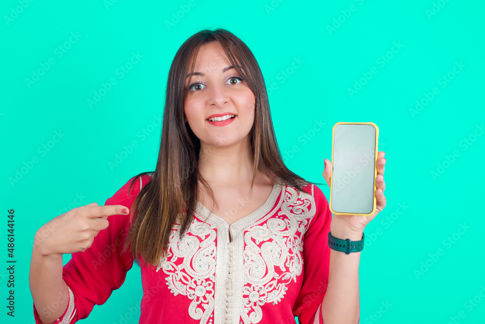 Attractive cheerful young beautiful moroccan woman wearing traditional caftan dress over green background holding in hands cell showing black screen Attractive cheerful young beautiful moroccan woman wearing traditional caftan dress over green background holding in hands cell showing black screen