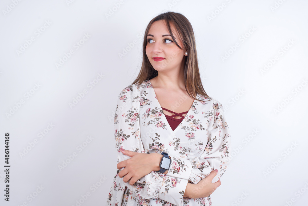 Pleased young Arab woman wearing floral dress over white background keeps hands crossed over chest looks happily aside Pleased young Arab woman wearing floral dress over white background keeps hands crossed over chest looks happily aside