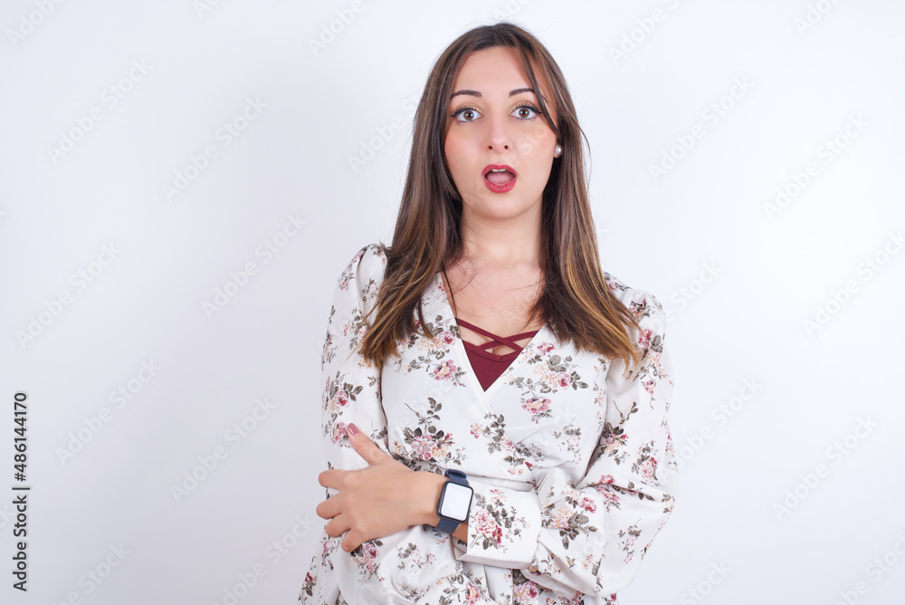 Shocked embarrassed young Arab woman wearing floral dress over white background keeps mouth widely opened. Hears unbelievable novelty stares in stupor Shocked embarrassed young Arab woman wearing floral dress over white background keeps mouth widely opened. Hears unbelievable novelty stares in stupor