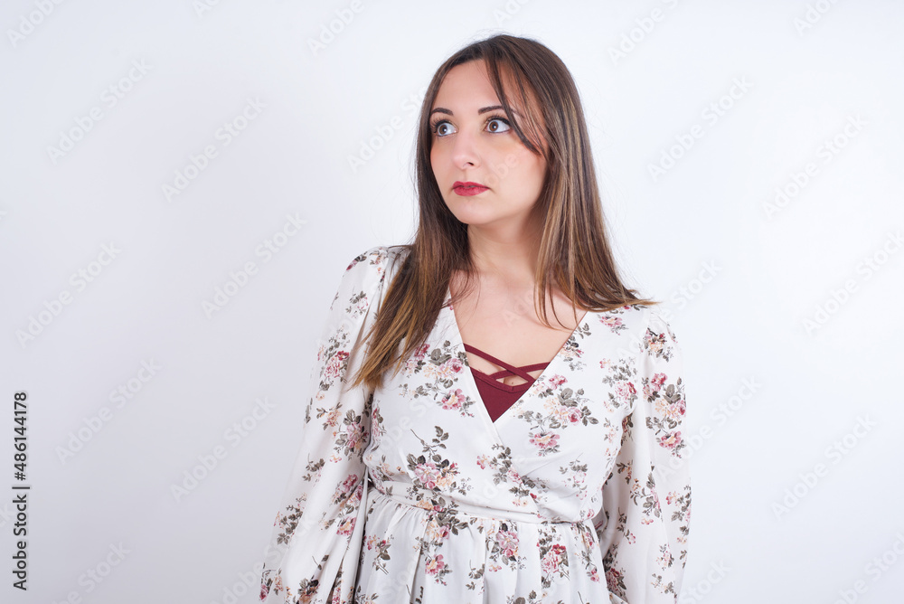 young Arab woman wearing floral dress over white background stares aside with wondered expression has speechless expression. Embarrassed model looks in surprise young Arab woman wearing floral dress over white background stares aside with wondered expression has speechless expression. Embarrassed model looks in surprise