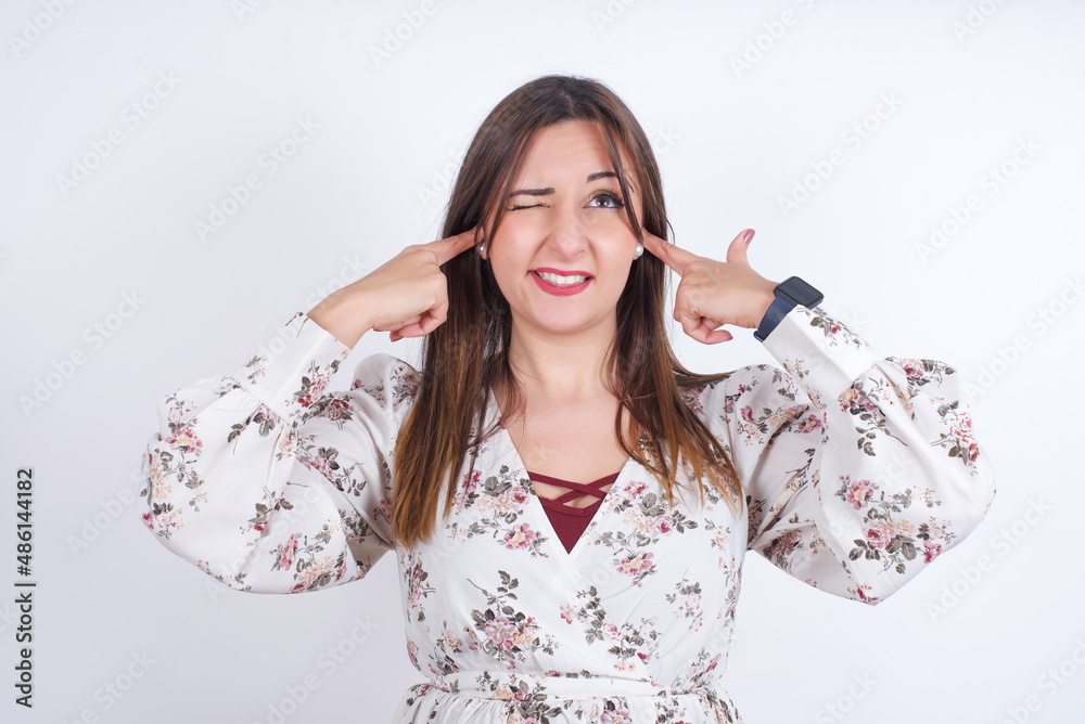 Happy young Arab woman wearing floral dress over white background ignores loud music and plugs ears with fingers asks to turn off sound Happy young Arab woman wearing floral dress over white background ignores loud music and plugs ears with fingers asks to turn off sound