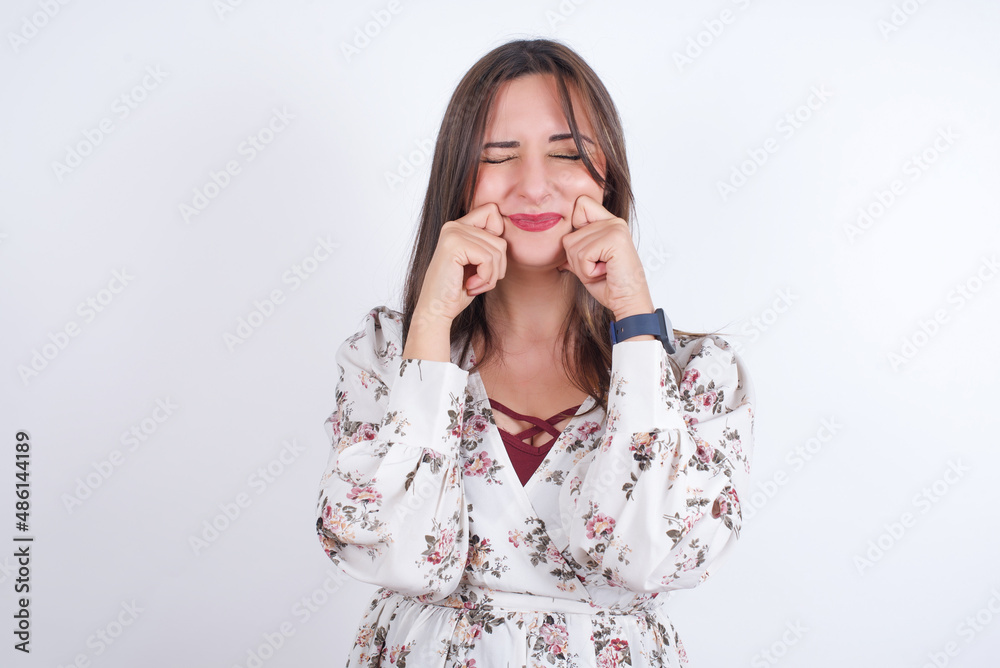 Pleased young Arab woman wearing floral dress over white background with closed eyes keeps hands near cheeks and smiles tenderly imagines something very pleasant Pleased young Arab woman wearing floral dress over white background with closed eyes keeps hands near cheeks and smiles tenderly imagines something very pleasant