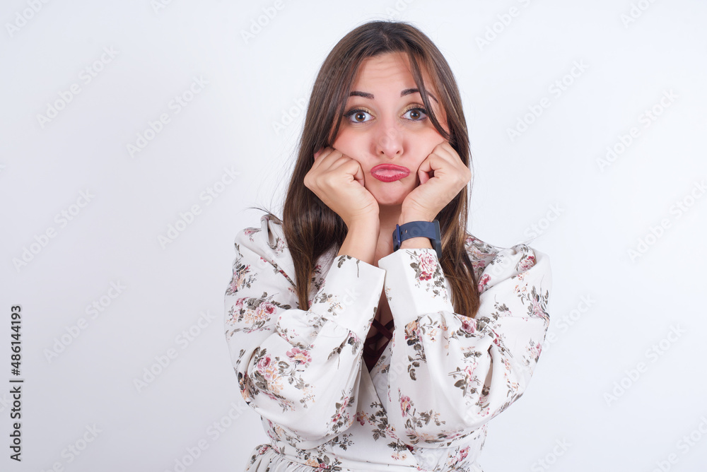 young Arab woman wearing floral dress over white background with surprised expression keeps hands under chin keeps lips folded makes funny grimace young Arab woman wearing floral dress over white background with surprised expression keeps hands under chin keeps lips folded makes funny grimace