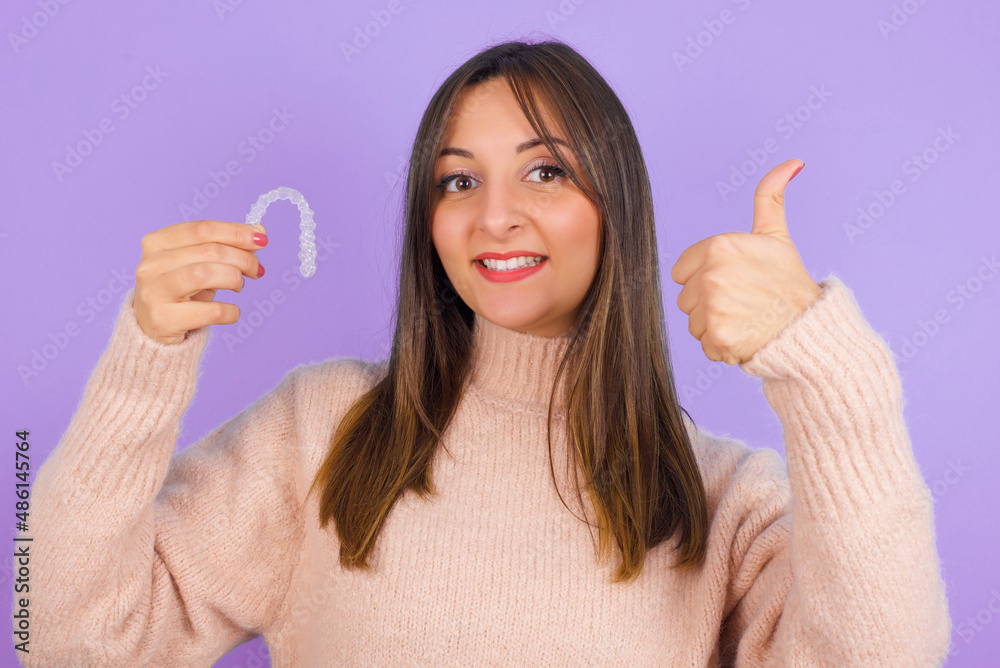 Young beautiful brunette woman standing against purple background holding an invisible retainer or braces and pointing with thumb up. 