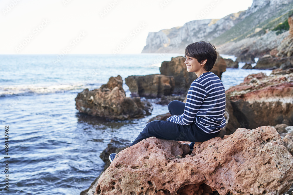 The boy is sitting on a rock by the sea.