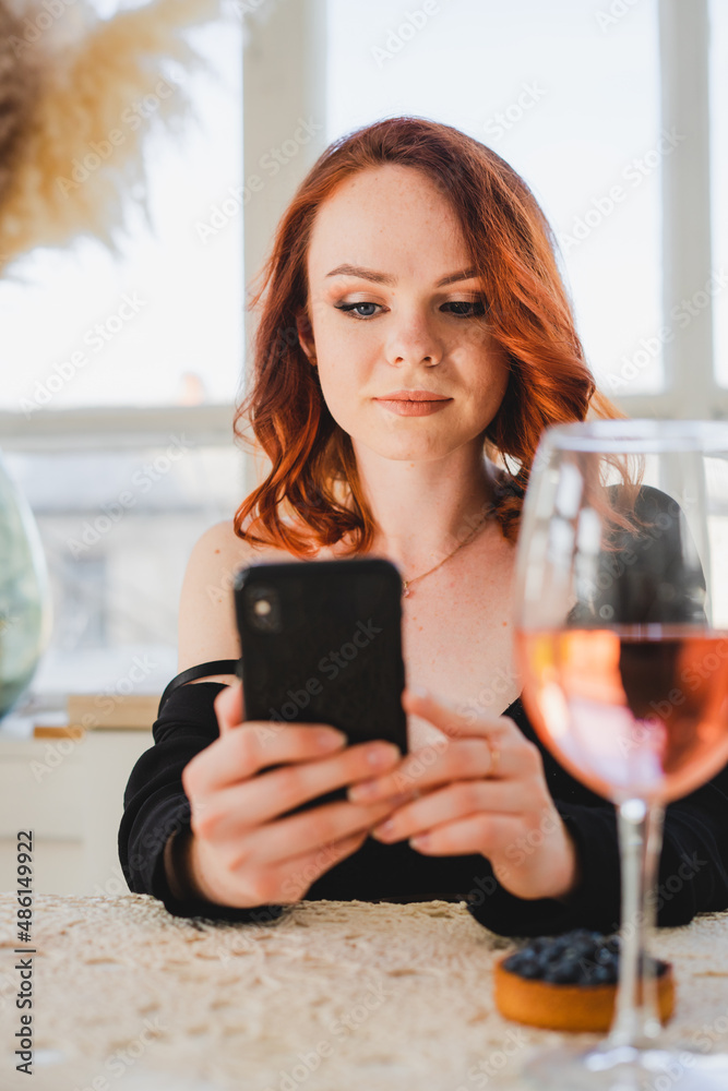 Beautiful girl with red hair holding a smartphone. There's a glass of rose wine on the table. The girl is resting. Beautiful girl with red hair holding a smartphone. There's a glass of rose wine on the table. The girl is resting.