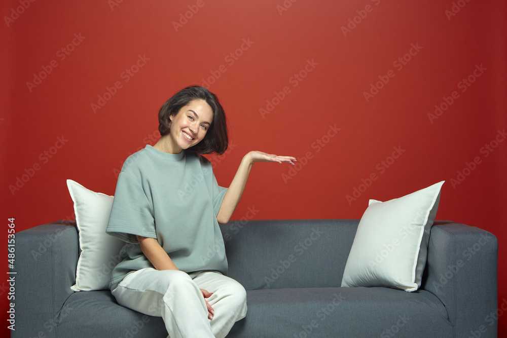 Happy young girl showing copy space by palm, advertise new product or service, sitting on sofa. Sale, online store offer Happy young girl showing copy space by palm, advertise new product or service, sitting on sofa. Sale, online store offer