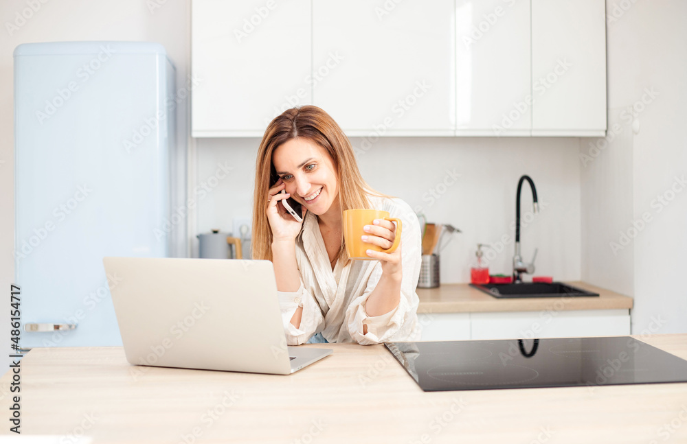 freelancer talking on mobile phone, drinking coffee and using laptop sitting at table in kitchen with modern interior. freelancer talking on mobile phone, drinking coffee and using laptop sitting at table in kitchen with modern interior.