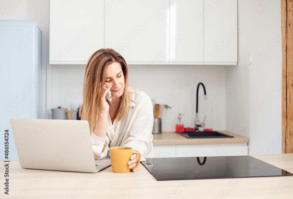 freelancer talking on mobile phone, drinking coffee and using laptop sitting at table in kitchen with modern interior. 
