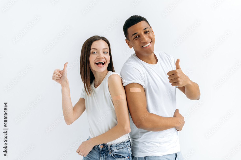 Joyful happy caucasian girl and hispanic guy, are got a vaccine, stand on a white background with medical plasters on their shoulders, look at the camera, smiling, showing thumb up gesture Joyful happy caucasian girl and hispanic guy, are got a vaccine, stand on a white background with medical plasters on their shoulders, look at the camera, smiling, showing thumb up gesture