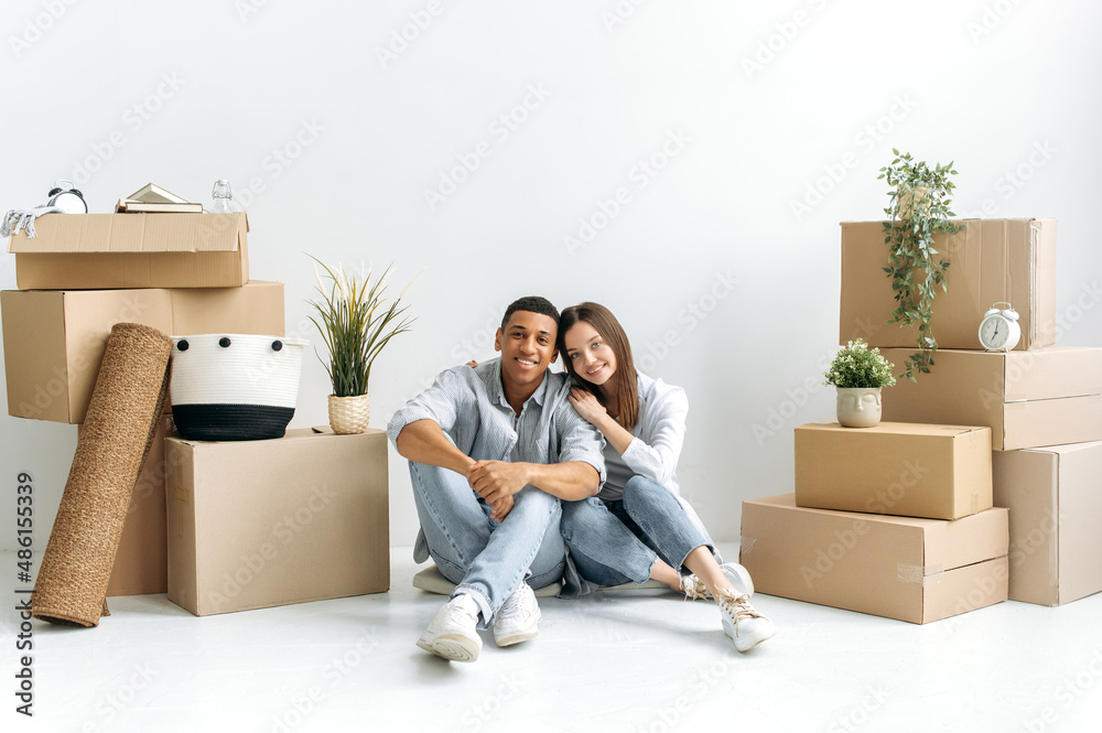 Happy in love couple, caucasian woman and hispanic man, stylishly dressed, sit in their first own house on the floor, there are card boxes with things around, they smiling, invested in real estate Happy in love couple, caucasian woman and hispanic man, stylishly dressed, sit in their first own house on the floor, there are card boxes with things around, they smiling, invested in real estate