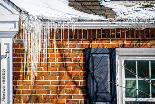 Large icicles hanging from snow covered roof of brick house indicating poor roof insulation.