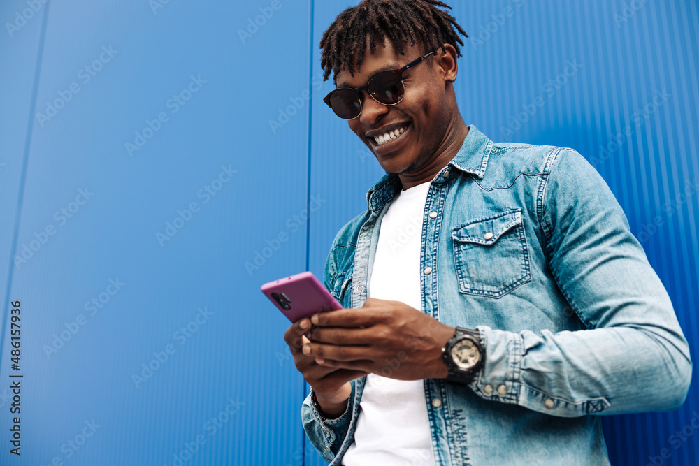 happy curly african american man wearing sunglasses using mobile phone and smiling against blue wall background