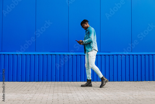 stylish African American man in full growth, uses a smartphone and walks past a blue wall while going to work © Shopping King Louie