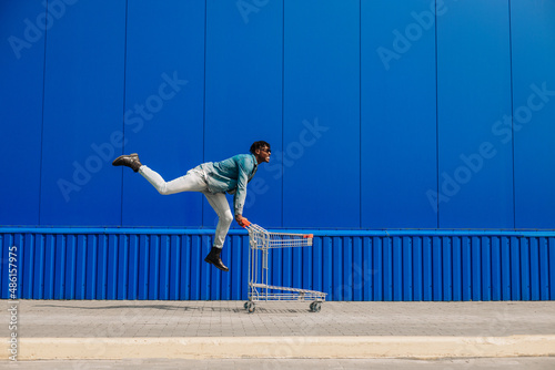 african american man in casual wear and sunglasses running with empty shopping cart outdoors against blue wall