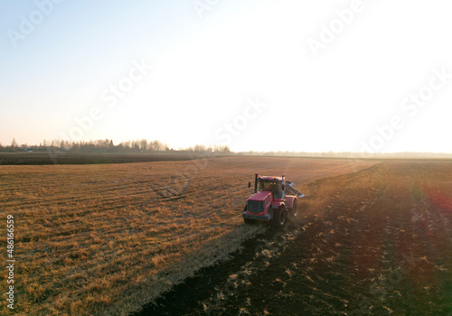 Tractor Plowing field on sunset. Red Tractor with Plough on Plowed. Ploughing and Soil Tillage. Agricultural Tractor on Cultivation Field for Sowing Seeds. Big Tractor During Field Cultivating. .