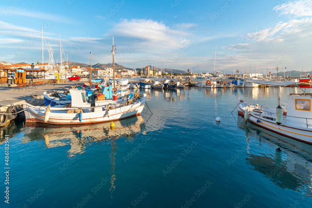Colorful fishing boats line the harbor of the Greek island of Aegina, Greece at dusk, with the waterfront promenade and village in view.	