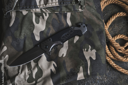 Tactical folding knife for survival and rope on camouflage military clothing.