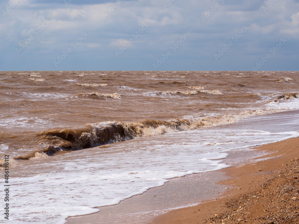 Chocolate-brown waves roll towards the sandy shore under the blue sky after a hurricane. Muddy sea water from strong wind waves. The natural state of the shallow Sea of Azov Chocolate-brown waves roll towards the sandy shore under the blue sky after a hurricane. Muddy sea water from strong wind waves. The natural state of the shallow Sea of Azov